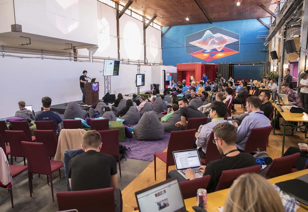 A large group of people attending a tech conference or workshop, with a speaker presenting on stage and attendees seated on chairs and beanbags, many working on laptops.