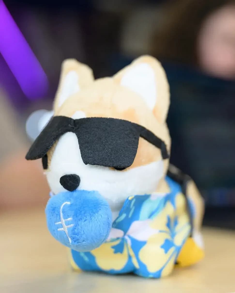 A cute plush toy of a corgi wearing sunglasses and a blue floral shirt, holding a blue ball with a filecoin logo embroidered on it.