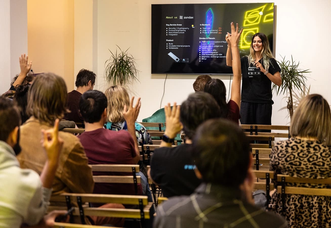 A woman gives a presentation to a seated audience, several participants raising their hands to ask questions in a modern meeting room.
